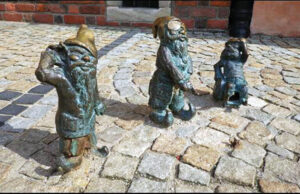 trzy krasnale z brązu ustawione na kostce brukowej, w tle ceglany budynek