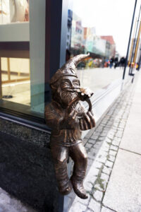 Widać krasnala siedzącego na kamiennym parapecie na tle witryny sklepowej.