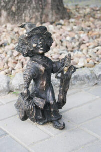 Krasnal z brązu idzie trzymając torby z zakupami i sukienkę na wieszaku.