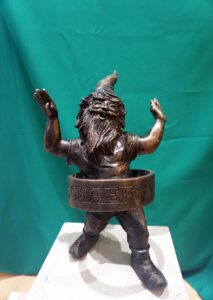 Krasnal z hula hopem stoi na postumencie na tle zielonej draperii.