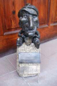 Krasnal z dużą jajowatą głową, siedzi na małym kamieniu, trzyma w dłoniach w dłoniach