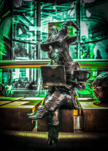 Figura z brązu przedstawia koziołka w nowoczesnej stylizacji, trzyma filiżankę kawy i laptopa, a na jego twarzy widoczne są ciemne okulary.