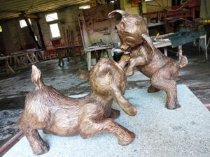 Dwie rzeźby koziołków o charakterze bajkowym, stoją w pozie rozbrykanej jakby bawiły się ze sobą. Na szarym granitowym postumencie, w tle widoczna pracownia kamieniarska.