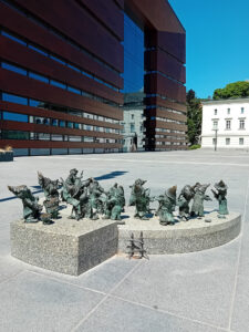 Grupa krasnali 21 wrocławskich grających na instrumentach muzycznych, ustawione na kamiennym postumencie na tle Narodowego Forum Muzyki