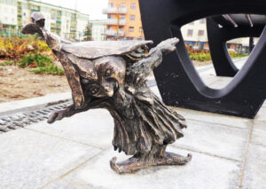 Na obrazku widać krasnala suwalskiego „Modraszka” – małą rzeźbę z brązu przedstawioną w dynamicznej, radosnej pozie. Postać jest ubrana w prostą sukienkę i spiczastą czapkę. Wzrok krasnala skierowany jest lekko w dół, ręce ma wyciągnięte na boki. Figura stoi na prostokątnej płycie chodnikowej. W tle widać fragment ławki i osiedla mieszkaniowego.