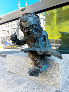 Krasnal z brązu siedzi na granitowym murku z otwartą książką i drapie się po głowie batutą.