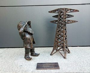 Krasnal Voltuś z uniesionymi rękami, zerka na stojący obok słup energetyczny, przed siedzibą Tauronu